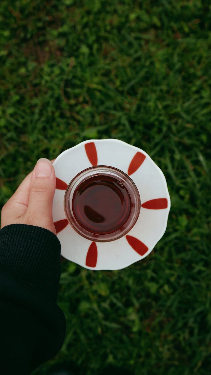 A person holding a traditional glass of tea above grassy ground outdoors.